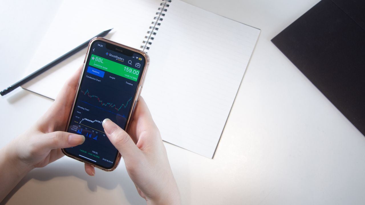 Close-up view of hands using a smartphone stock market app, with a notebook and pencil resting on a desk.